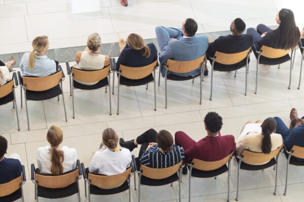 Présentateur en costume parlant à un auditoire assis en demi-cercle dans une salle lumineuse, contexte leadership stratégique et communication en groupe.