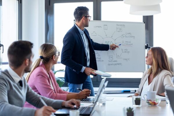 Salle de réunion avec un homme en blazer présentant un diagramme sur un tableau blanc, devant un groupe en discussion. pilotage stratégique entreprise pme intégré.