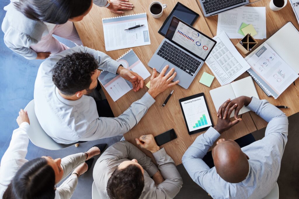 Réunion de travail vue du dessus : plusieurs professionnels autour d’une table avec ordinateur portable, tablettes et documents, discutant et échangeant des données. Alt text adapté au contexte « soft skills manager de demain ».