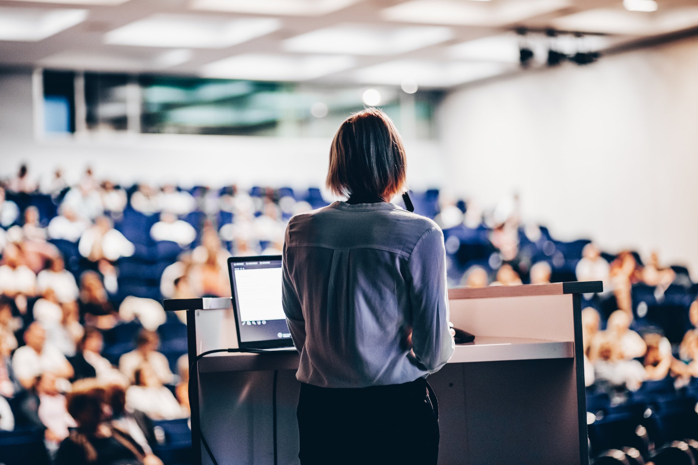 Une présentatrice debout derrière un pupitre devant une salle pleine d’auditeurs.
